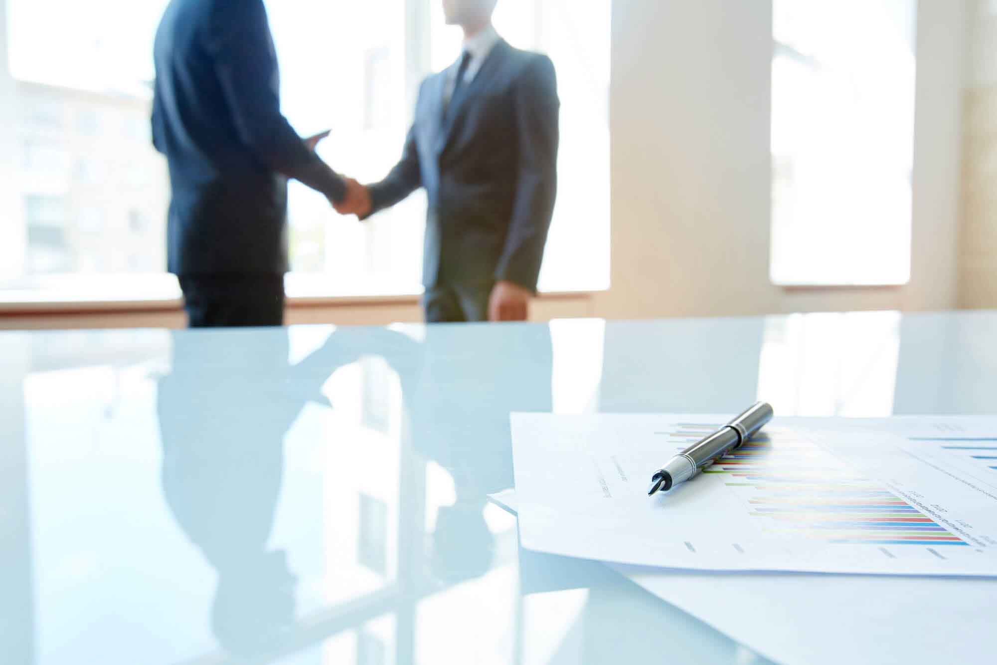 image of documents and pen on the table with 2 professionals shaking hands in background